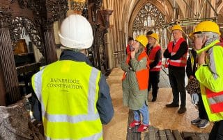 A group of people wearing hi-viz jackets and hard hats stand on scaffolding facing an ornate organ case, listening to a person wearing a vest that reads 'Bainbridge conservation'