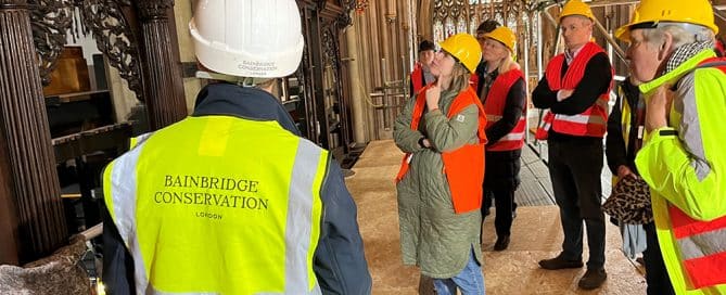 A group of people wearing hi-viz jackets and hard hats stand on scaffolding facing an ornate organ case, listening to a person wearing a vest that reads 'Bainbridge conservation'