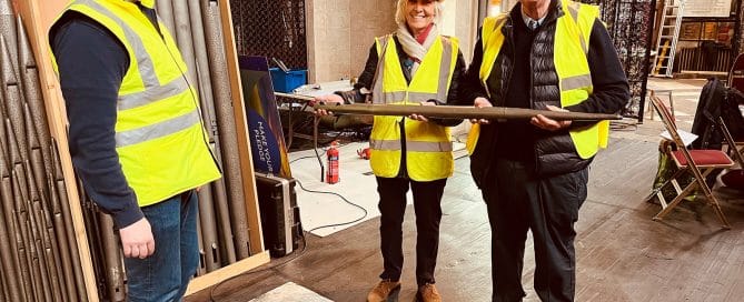 Three people wearing hi-vis jackets. Two are smiling and holding an organ pipe