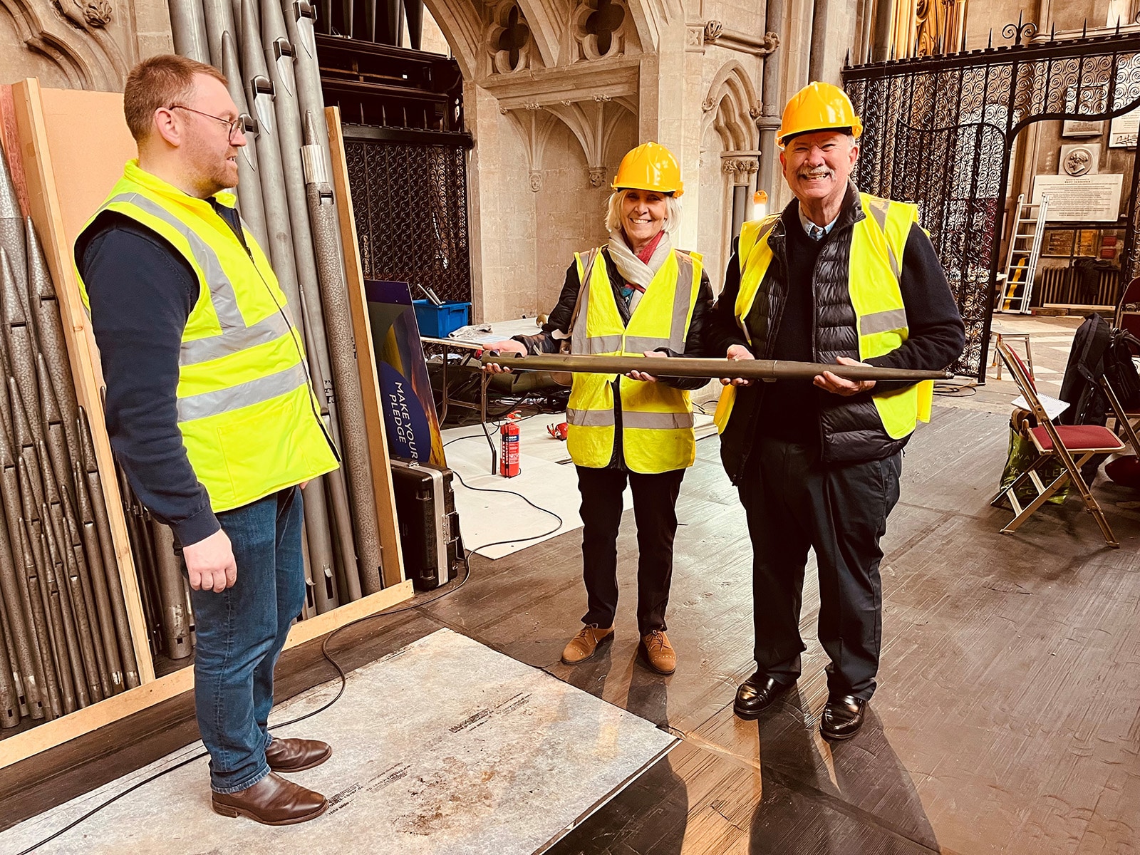 Three people wearing hi-vis jackets. Two are smiling and holding an organ pipe
