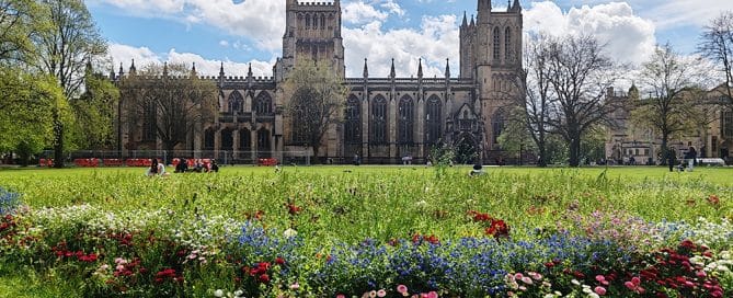 Bristol Cathedral on a bright spring day, with a green in front and a bed of spring flowers in the foreground.